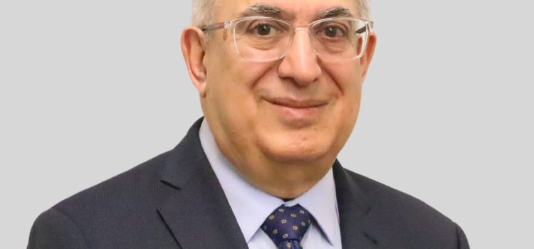 Portrait of a middle-aged man with gray hair and glasses, wearing a dark suit and patterned purple tie against a light gray background.