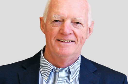 Professional headshot of an older man in a navy blazer and striped shirt, smiling against a light gray background.