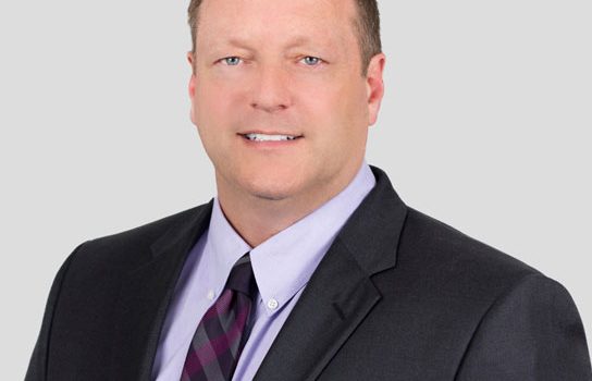 Professional head-and-shoulders portrait of a man in a dark suit with a lavender shirt and striped tie against a light gray background.