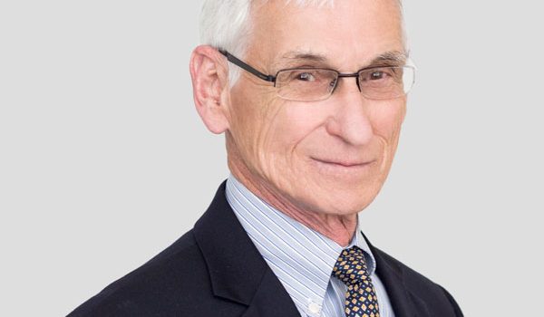 Professional headshot of an older man with white hair and glasses, wearing a dark suit and striped shirt.