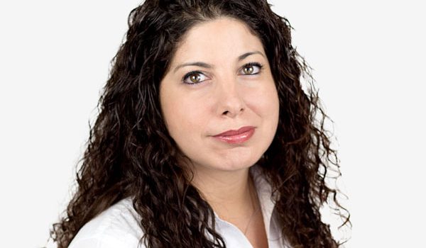 Portrait of a woman with long dark curly hair wearing a white shirt, looking at the camera against a white background.