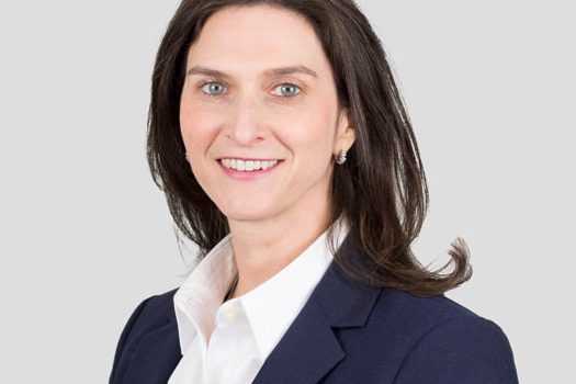 Professional headshot of a woman in a navy blazer and white shirt, smiling at the camera in front of a light gray background.