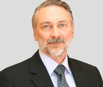 Professional portrait of a middle-aged man with gray hair and beard, wearing a dark suit and blue patterned tie against a light gray background