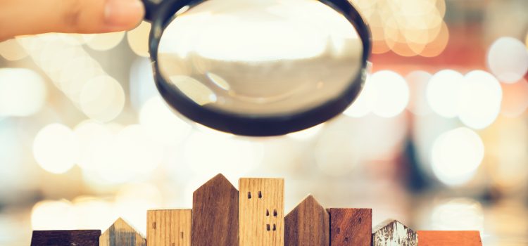 Hand holds a magnifying glass over a row of wooden toy houses, with blurred city lights in the background