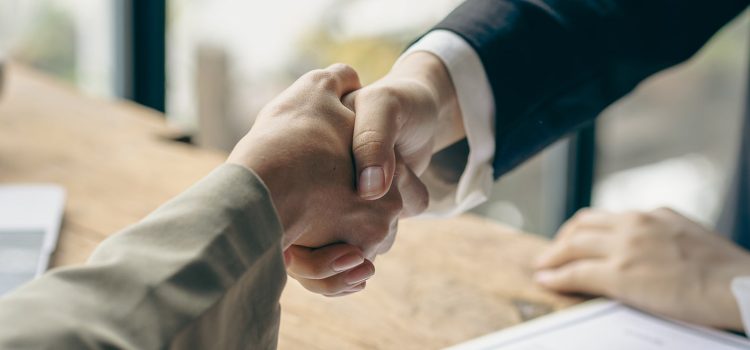 Two people shaking hands over a signed contract on a wooden table with documents nearby.