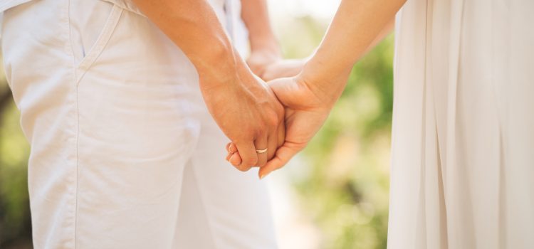 Couple holding hands outdoors, both wearing light clothing, wedding rings visible on the ring fingers.