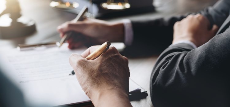 Close-up of a person writing with a gold pen on documents at a desk, scales of justice blurred in the background.