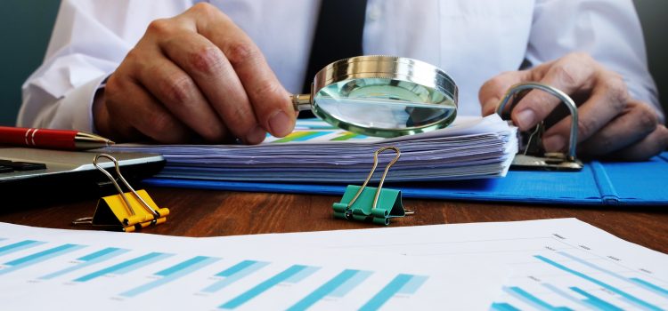 Person in a white shirt and black tie examines documents with a magnifying glass on a desk full of charts and clips.