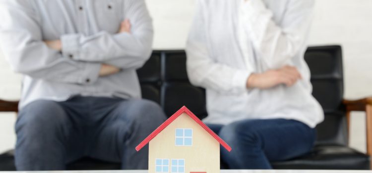 Small wooden toy house on a table with two blurred adults sitting in the background in a living room setting.
