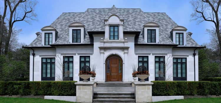 White mansion with a gray slate roof, central arched wooden door, and stone steps flanked by urn planters with plants.