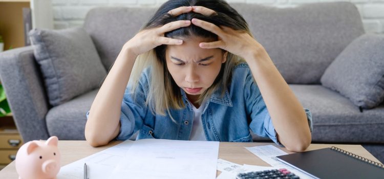 Woman sits at a desk with scattered papers, head in hands in a frustrated pose while balancing a calculator and notebook nearby.
