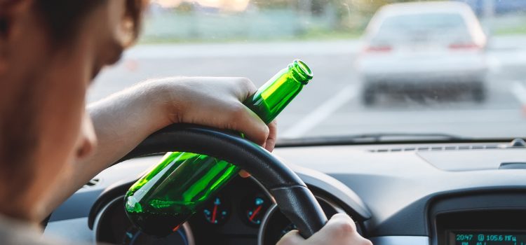 Driver holding a green bottle while steering a car, with a blurred road scenery outside the windshield.