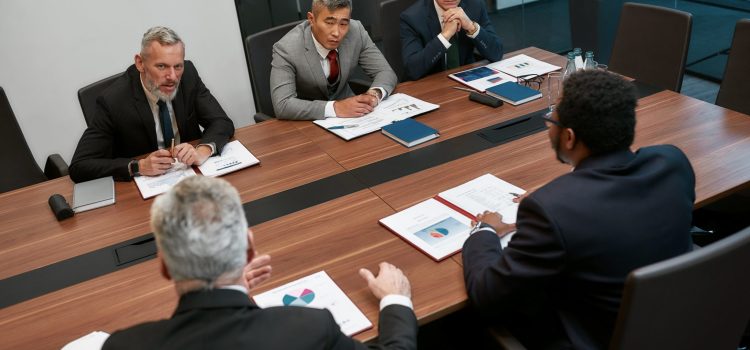 Five business professionals sit around a wooden conference table reviewing charts and documents during a meeting in a glass-walled office.