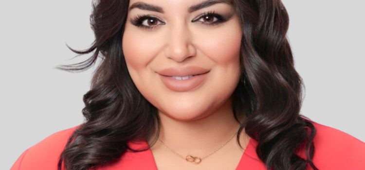 Close-up portrait of a smiling woman with dark wavy hair, wearing a coral blouse and subtle makeup, against a gray background.