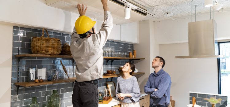 Man wearing a yellow hard hat reaches up to adjust a ceiling air vent while two coworkers watch in a modern kitchen showroom. The scene shows HVAC work in progress.