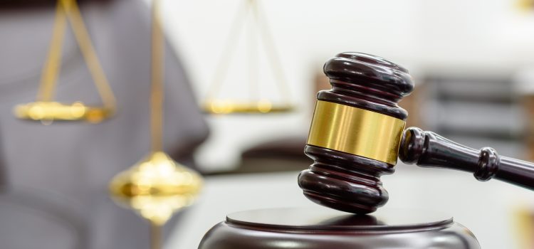 Close-up of a judge's gavel resting on a sound block with blurred scales of justice in the background, symbolizing law.