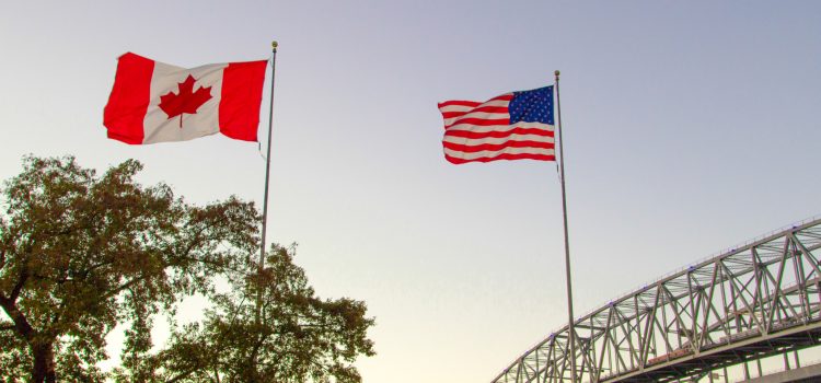 Canada and United States flags flying on flagpoles over a treed area, with a large steel bridge visible on the right against a clear sky.