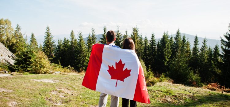 Two people stand with a Canadian flag draped over their backs in a sunny, forested mountain area (back view). TODO