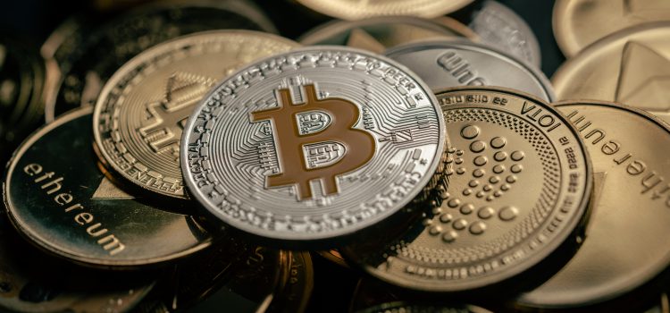 Close-up of a silver Bitcoin coin with an orange B logo, surrounded by other gold coins on a dark surface.