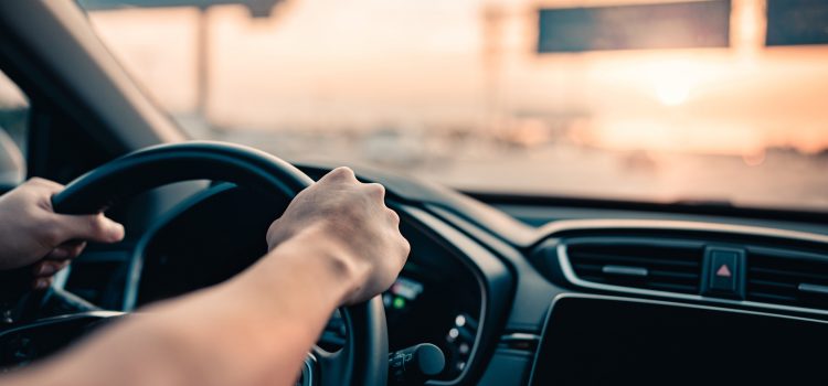 Hands on steering wheel as the car moves at dusk with a blurred highway in the background.0-0