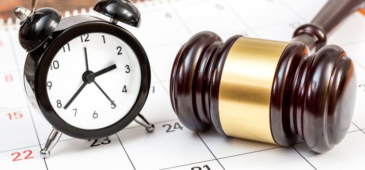 Analog black alarm clock and a wooden gavel resting on a calendar page, suggesting a deadline or time-related law theme.