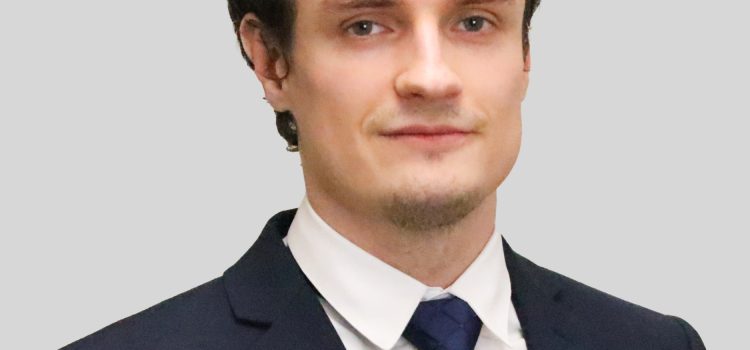 Close-up portrait of a man in a dark suit, white shirt, and navy tie against a light gray background.