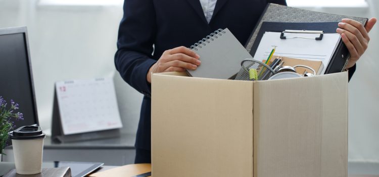Person in a dark blazer packs office items into a large cardboard box, holding notebooks and folders amid desk supplies.