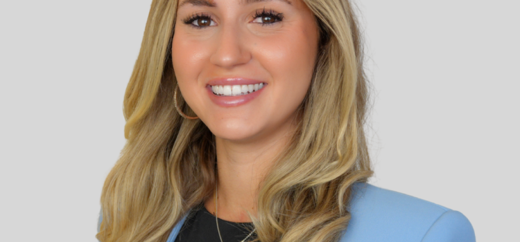Smiling woman with long blonde hair in a light blue blazer, black top, against a gray background.