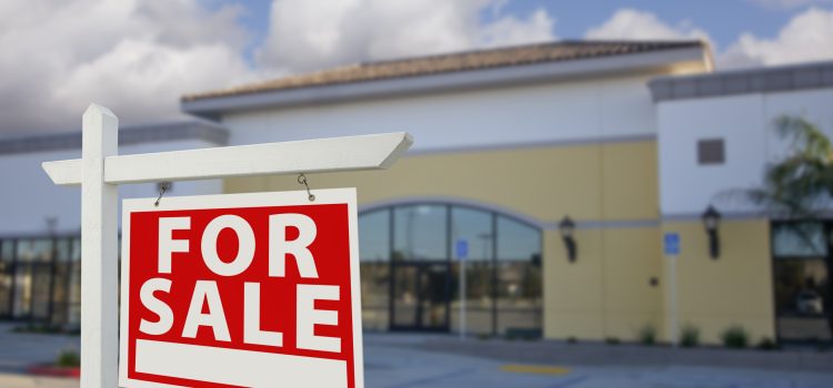 Red and white 'FOR SALE' sign in foreground outside a shopping plaza against a blue sky with clouds.