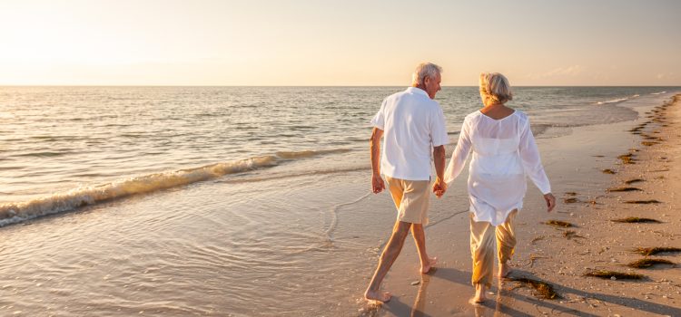 Elderly couple walking barefoot along the sunny shoreline, holding hands at sunset.