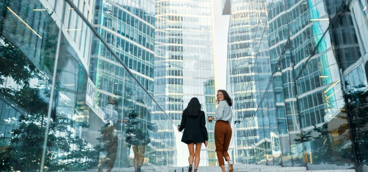 Two women in business attire walk up a stone staircase between tall glass skyscrapers, smiling at each other as they ascend.