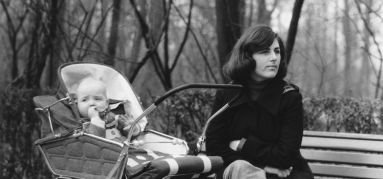 Woman sits on a park bench with a baby in a vintage stroller beside her, both facing left, black-and-white photo.