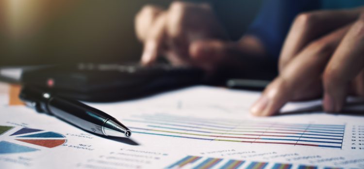 Close-up of a pen on colorful bar and line charts on financial documents, with blurred hands in the background suggesting analysis.