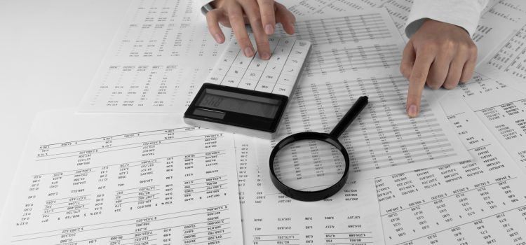 Hands typing on a calculator surrounded by financial documents and a magnifying glass.