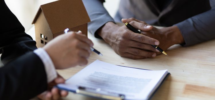Two people sign documents at a table, with a small cardboard house model beside them as part of a real estate or mortgage discussion.