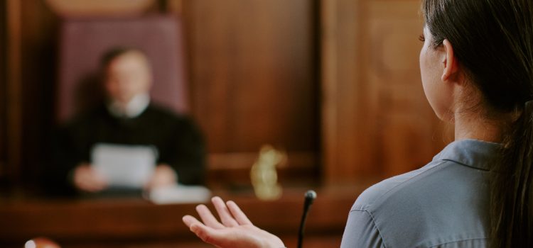 Person facing away, gesturing with hands while speaking in a courtroom; judge blurred in the background.