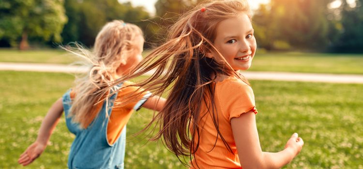 Two girls run through a sunlit park, smiling as their hair blows in the breeze.