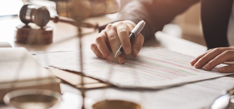 Close-up of a hand signing documents with a pen, pages showing colored highlights on a desk; a gavel and glasses blur in the background.