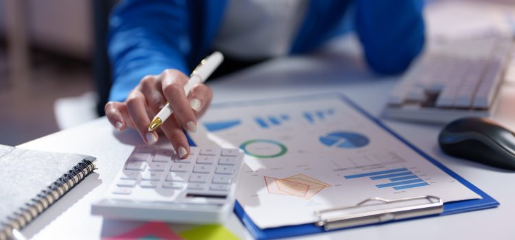 Person in a blue sweater using a white calculator while reviewing colorful charts and graphs on a desk.
