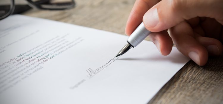 Close-up of a hand signing a document with a silver pen on a wooden desk, glasses blurred in the background.