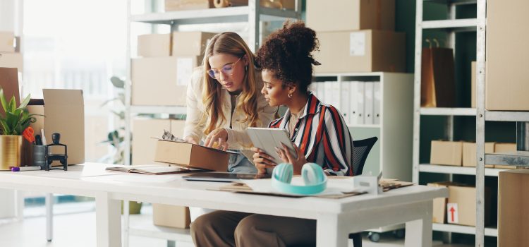 Two women collaborate at a work desk in a storage room, one showing a clipboard while the other holds a tablet as they review packaging items.