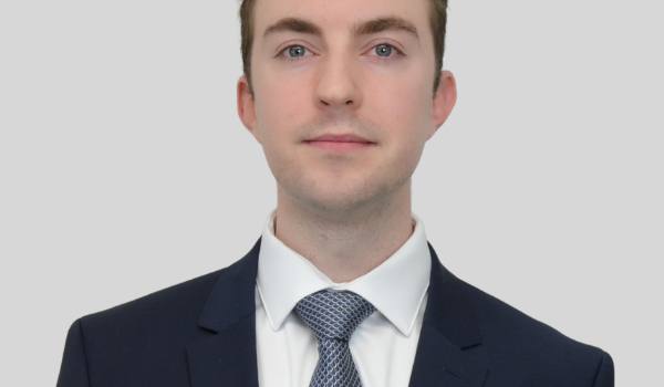 Professional headshot of a young man in a navy suit and blue patterned tie against a light gray background.