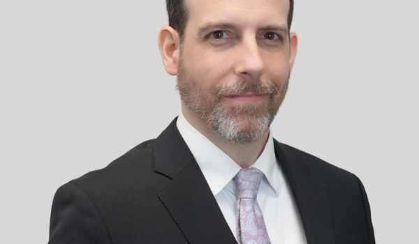 Portrait of a man in a dark suit and light pink paisley tie, looking at the camera against a light grey background.