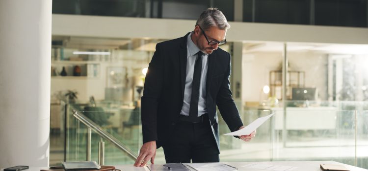 Professional man in a dark suit and glasses leans over a desk, reviewing documents in a modern office.