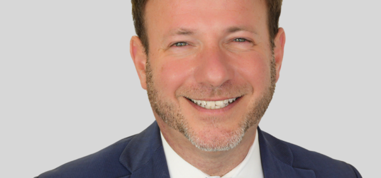 Head-and-shoulders portrait of a smiling man in a navy suit and blue tie against a light gray background, professional headshot.